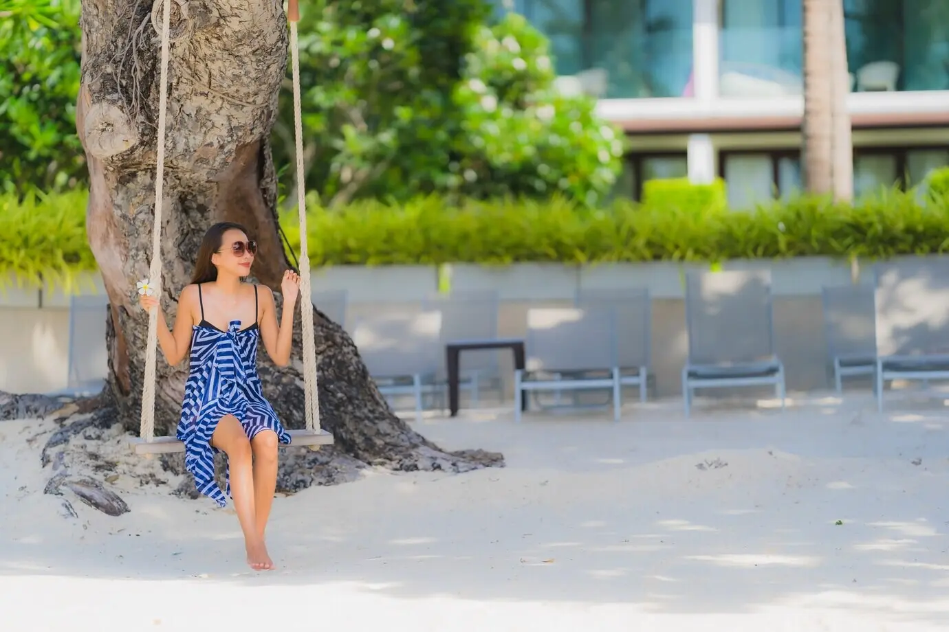 Porträt einer jungen asiatischen Frau, die auf einer Seilschaukel sitzt, umgeben von Meer, Strand, Ozean und einer Kokospalme.