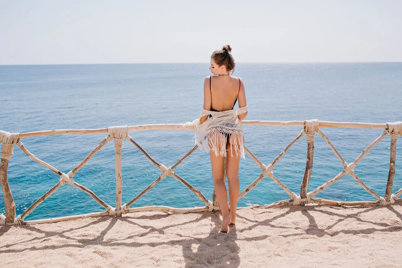 Schlankes, gebräuntes Mädchen mit niedlicher Frisur in Strickkleidung steht auf einer Terrasse und blickt auf das Meer. Beeindruckende junge Frau genießt im Sommerurlaub den Ozeanblick und posiert vor dem Wasser.